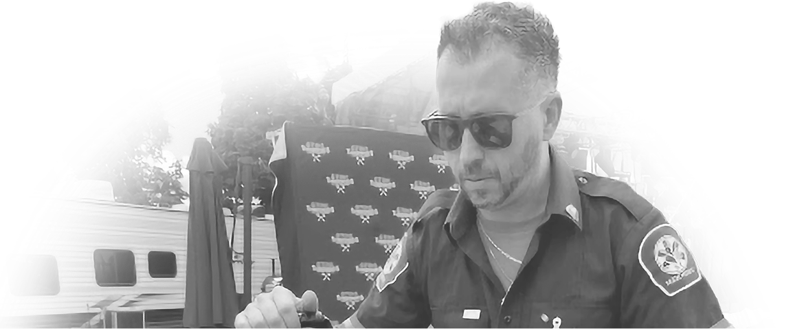 Man in sunglasses wearing a dark shirt with patches, possibly a uniform, in front of a backdrop with logos. Mood is serious and professional.