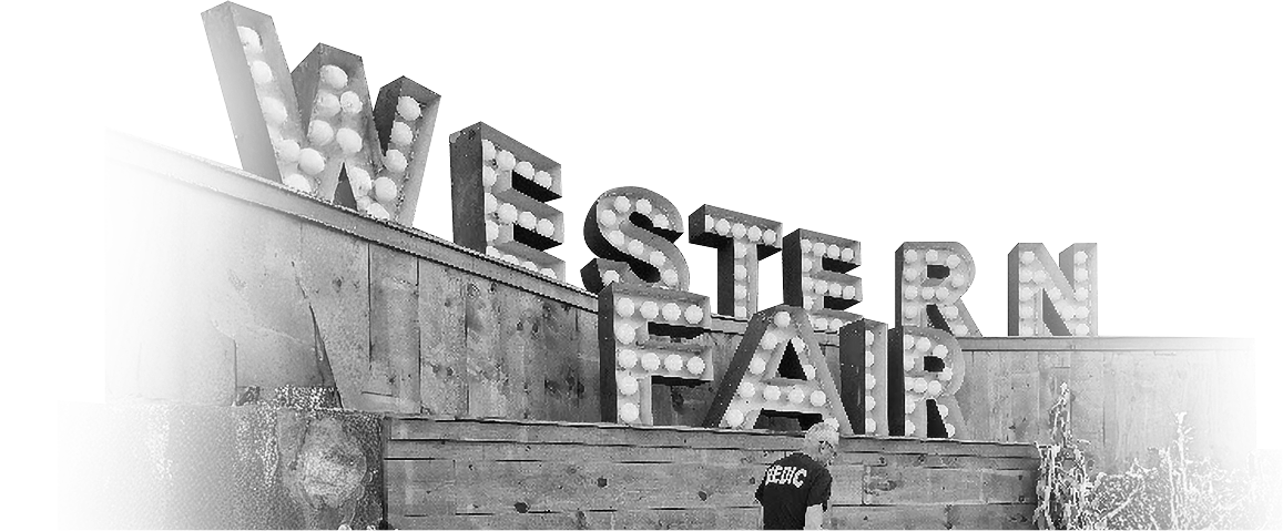 Illuminated "Western Fair" sign with vintage bulbs on a wooden backdrop. A person in a "MERC" shirt stands in the foreground. Classic fairground ambiance.