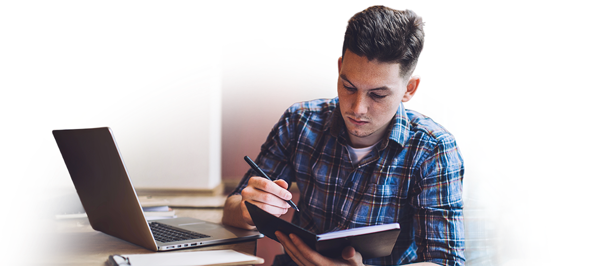 A student holding a notebook and a pen with his laptop open in front of him on a desk.