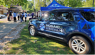 Blue Mosaic Medical SUV parked on grass near a sidewalk, with people gathering under a blue canopy in a park setting. Bright, sunny day.