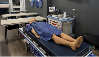 A medical training room featuring a mannequin on a stretcher, covered with a blue sheet. Equipment and a monitor display abstract visuals nearby.