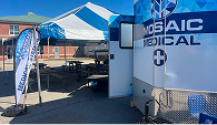 Outdoor medical setup with blue and white tent labeled "Mosaic Medical." Nearby is a banner beside the tent, and tables are visible underneath.