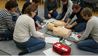 A group of people practicing CPR on a manikin, surrounded by instruction sheets and a red first aid kit, conveying a sense of focus and cooperation.