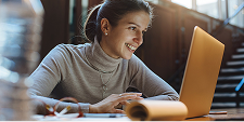 A woman in a cozy turtleneck works on a laptop with a focused smile. Warm sunlight streams through large windows, creating a relaxed, productive atmosphere.