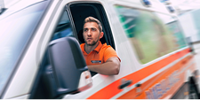 A man dressed in an orange Mosaic Medical shirt drives an ambulance down the street.