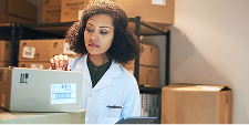 A woman in a lab coat examines a labeled box in a storage room filled with stacked cartons. She appears focused and organized, suggesting inventory work.