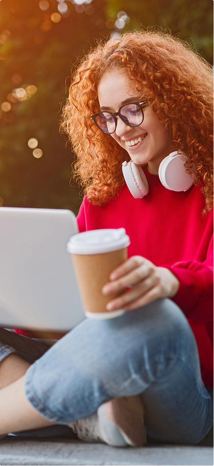 A smiling woman with curly red hair, wearing glasses and headphones, sits outdoors with a laptop and coffee cup. They wear a red sweater and jeans.