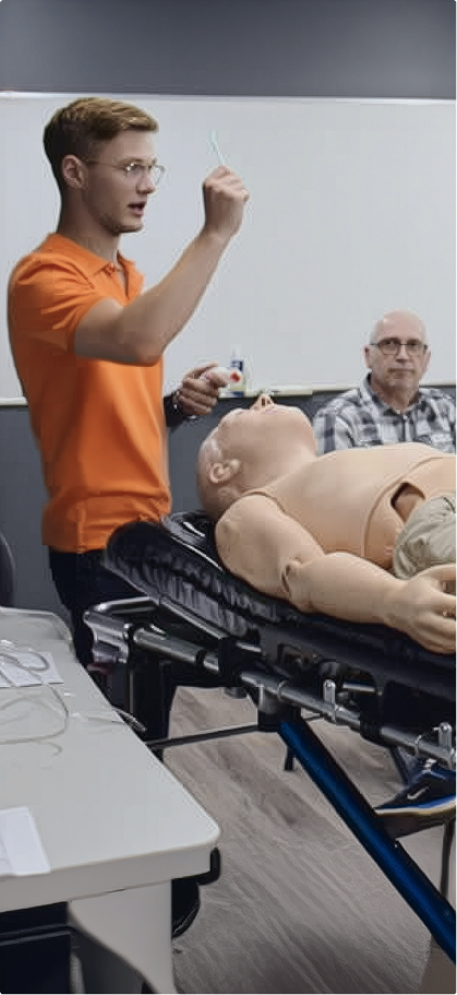 An instructor in an orange shirt demonstrates a medical procedure using a mannequin on a stretcher. A person watches attentively in the background.